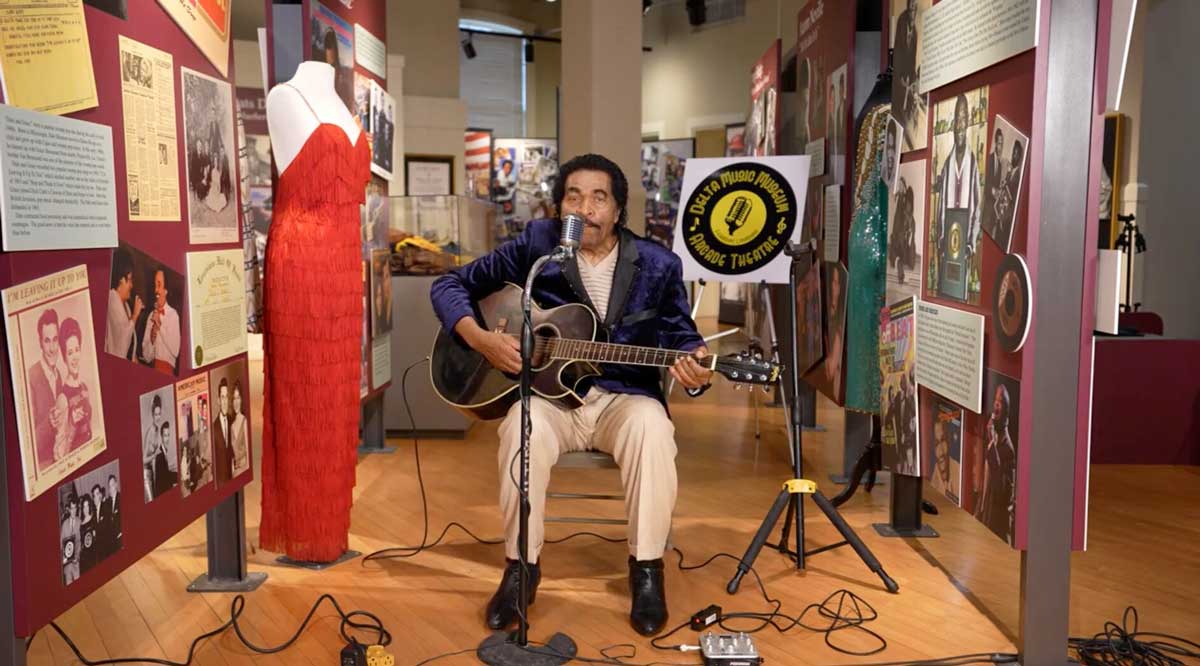 Bobby Rush is seated, playing guitar in front of a microphone in a museum. He's next to a display with a striking red ballgown and other music memorabilia