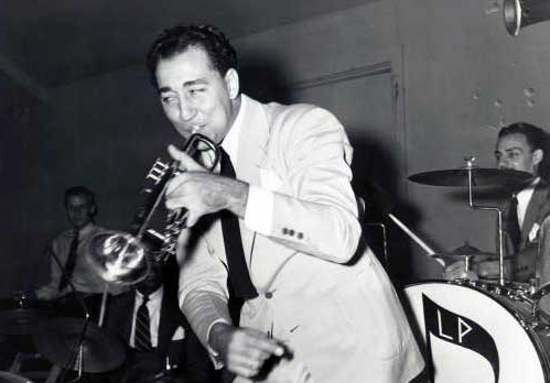 A trumpet-holding Louis Prima stands on stage with a band behind him. He wears a white suit with a black tie.