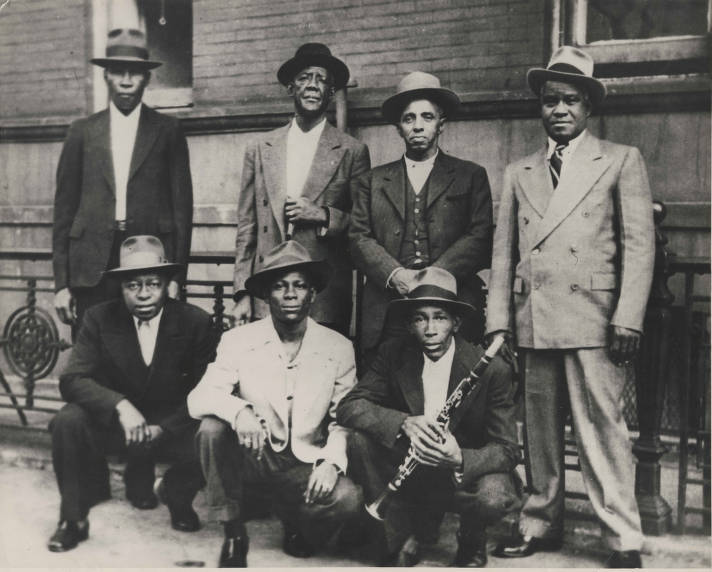 A black-and-white image of four standing and three squatting men posing for the camera on a street; one holds a clarinet