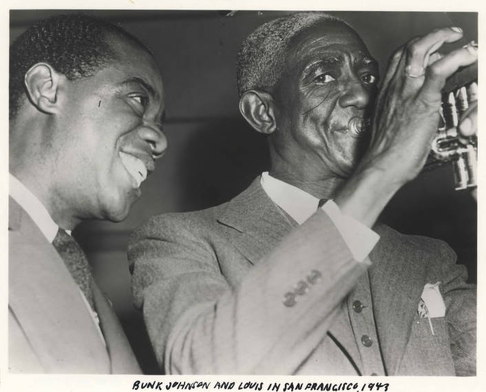 A smiling Louis Armstrong stands close to Bunk, who's playing his trumpet