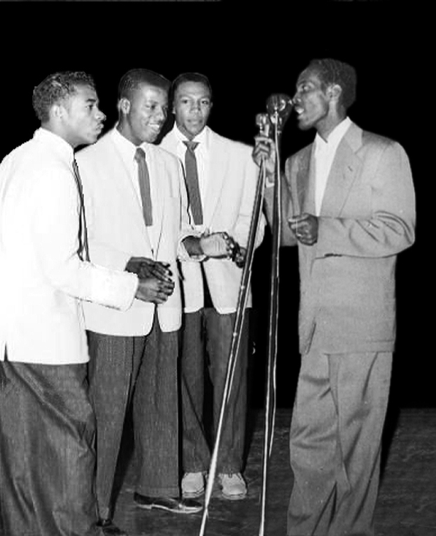 Three men in white jackets stand across from a dark-suited Percy as he sings into the mic