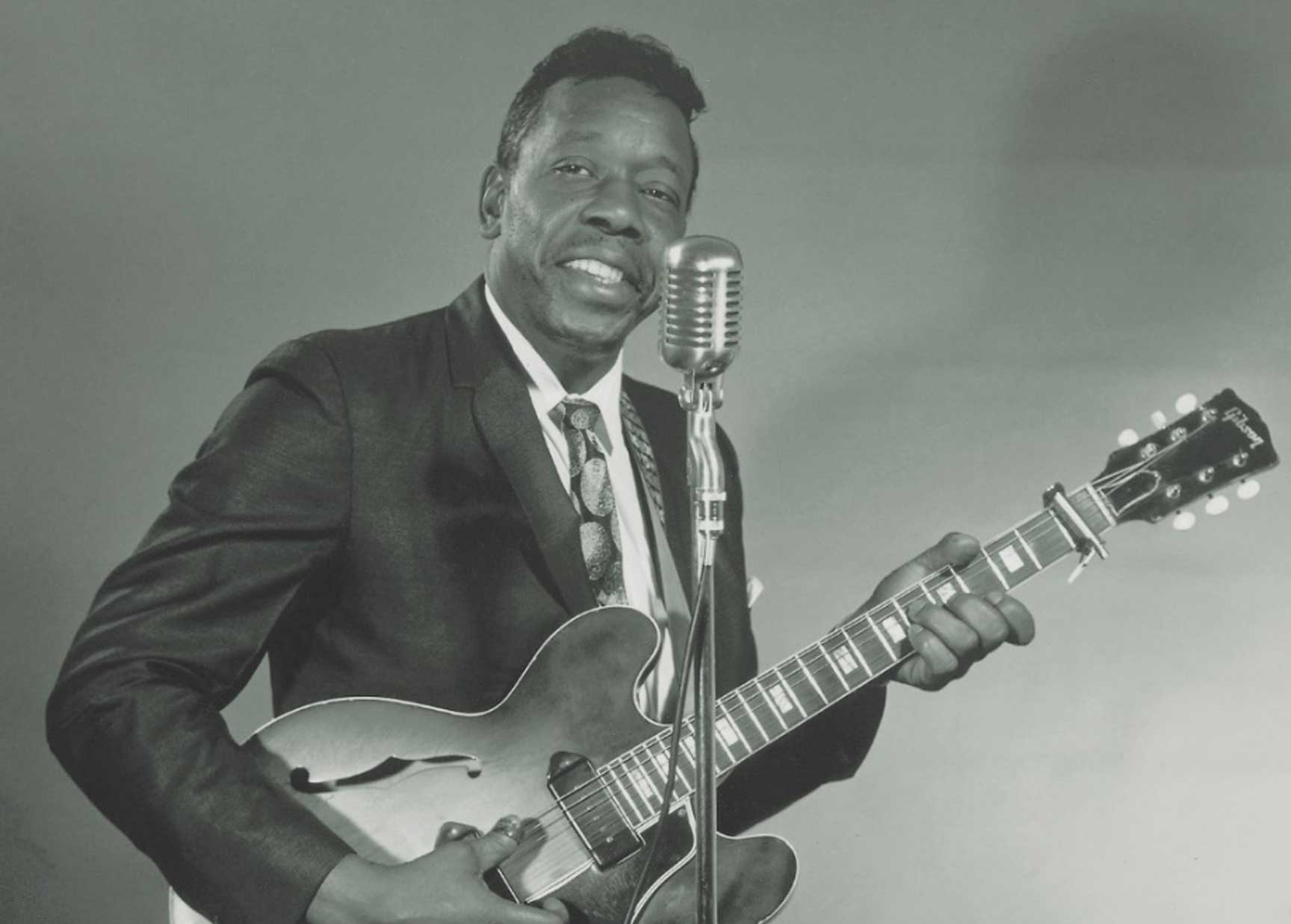 Slim stands jauntily behind a mic while holding his guitar