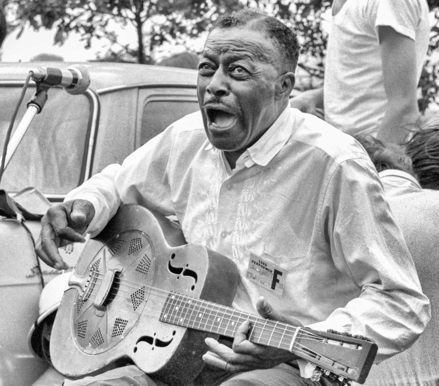 An older Son House sits on a chart and plays his guitar into a mic. He's outside, sitting next to a classic car and appears to be laughing