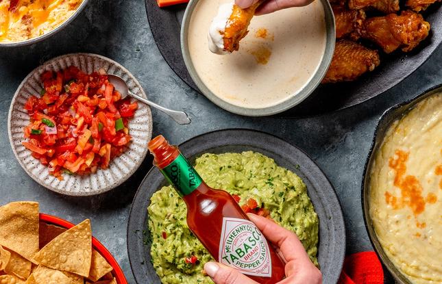 A Tabasco bottle is held over plates of sauces and other condiments, including guacamole and salsa.