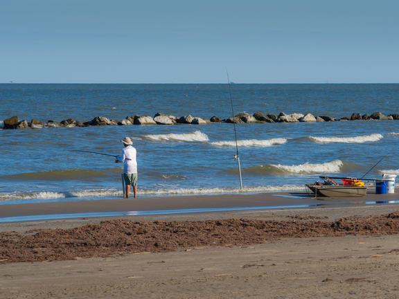 A man stands at the edge of the water holding a fishing rod at Grand Isle State Park, while his boat and other equipment sit just beyond the water.