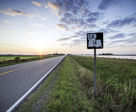 A rural two-lane highway, part of Louisiana's Cajun Corridor Byway, stretches into the distance. In the background the sun peeks over the horizon, brightening a cloudy sky.