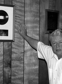 J.D. Miller standing proudly next to the gold record of Kitty Wells's "It Wasn't God Who Made Honky Tonk Angels." Wikimedia. Photo by John Broven. 