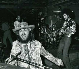 Dr. John sits at a piano singing enthusiastically while a man plays guitar behind him