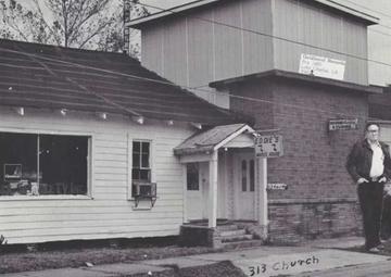 A man stands in front of two buildings, one a white clapboard house with a sign that says "Eddie's Music House," and one that's a double story, with a sign that says "Goldband Records."