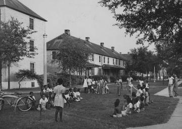 A black and white image of two housing blocks. On the lawn two dozen people lounge.