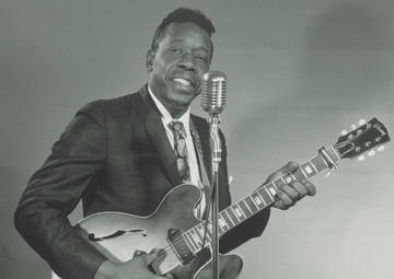 Slim stands jauntily behind a mic while holding his guitar