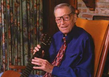 A color photo of Jimmie holding a guitar while sitting in a rocking chair. Curtains and a brick wall are behind him. He wears a navy dress shirt and patterned tie